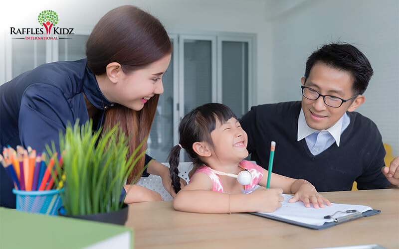 Smiling girl writing on clipboard at a table with two adults in a bright learning space.