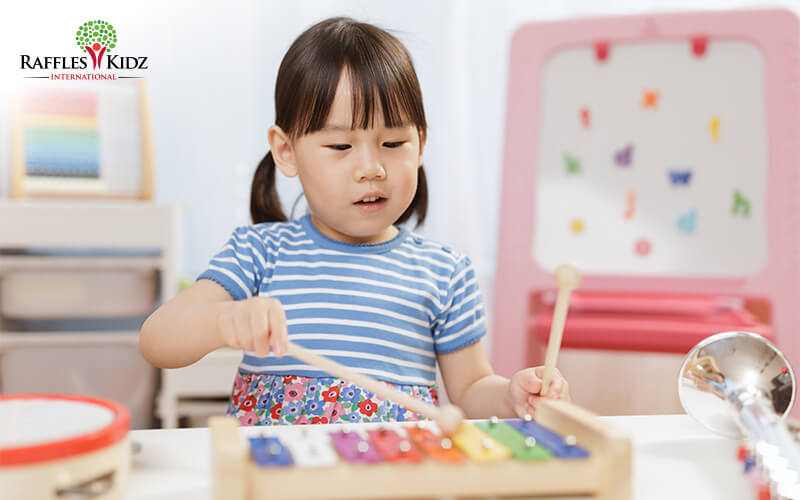 A young child plays a colorful wooden xylophone, holding two mallets and concentrating as she taps the keys in a bright, child-friendly room with toys and learning materials in the background.