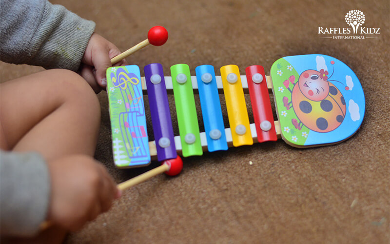 Child playing a colorful toy xylophone with wooden mallets on a carpeted surface.