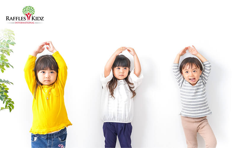 Three young children stand side by side against a white background, each raising their arms above their head in playful poses, wearing casual, colorful outfits and showing different facial expressions.