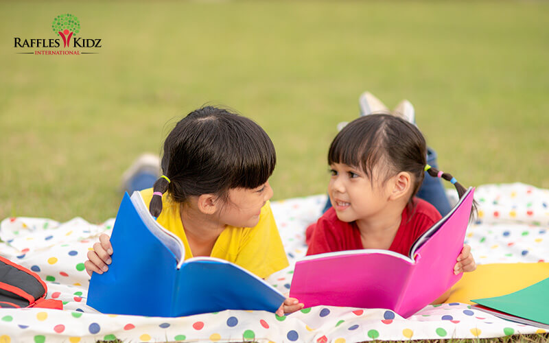 Two children read books on colorful blanket outdoors.