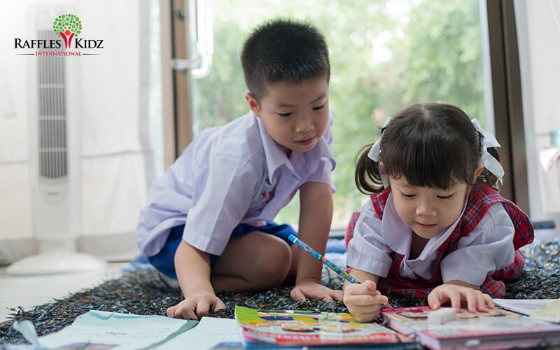 Two children in uniforms study workbook together on floor.