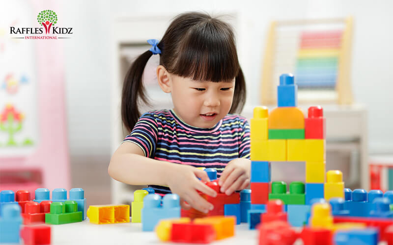 A young child with pigtails plays at a table, stacking colorful plastic building blocks to form a small structure in a bright classroom.