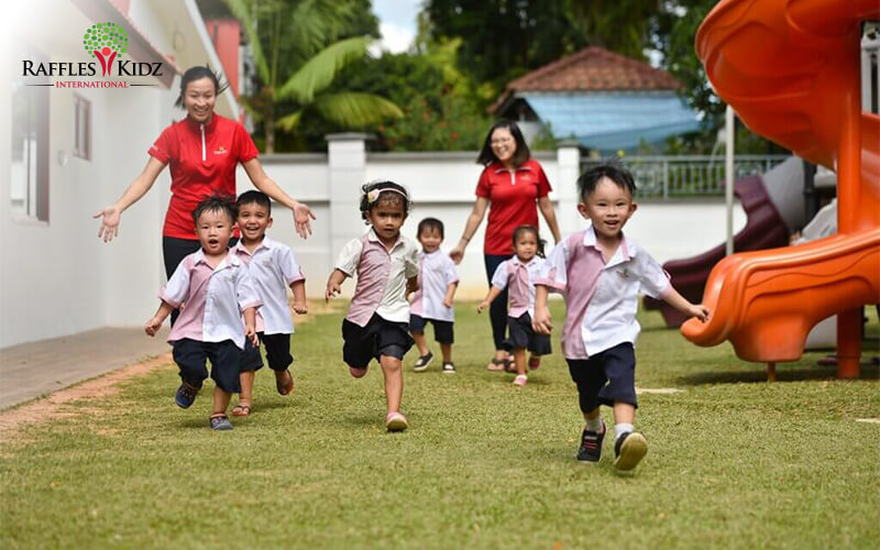 Group of children in matching uniforms running on grass near playground slide, supervised by two adults.