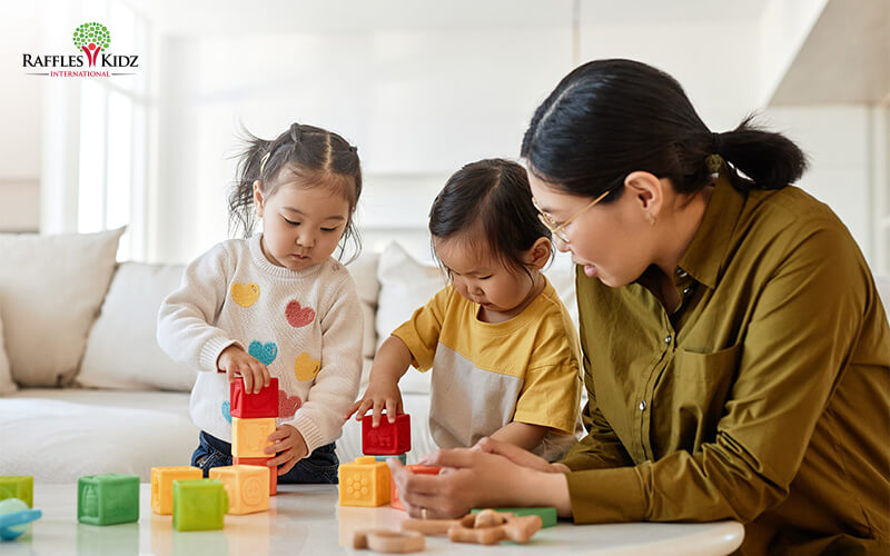 Raffles Kidz teacher guiding toddlers stacking colourful building blocks.