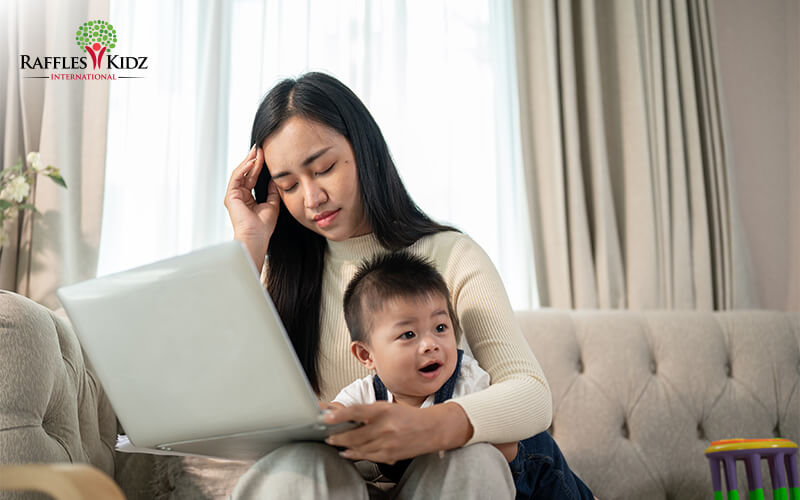 Tired Asian Woman sitting on a sofa using a laptop while holding a baby on her lap